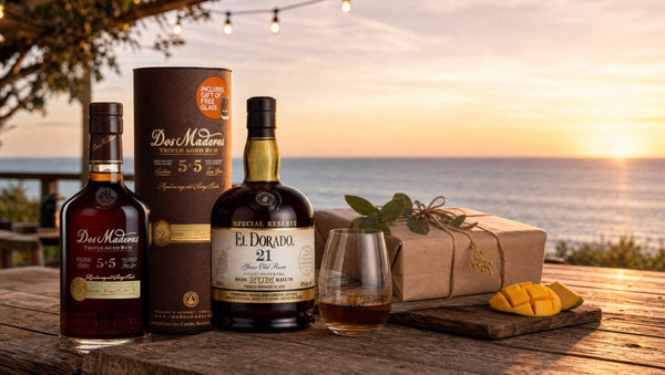 Dos Maderas 5+5 rum with its gift tube and El Dorado 21 Year Old rum on a beach deck at sunset, with a wrapped present